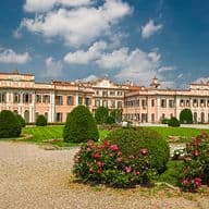 A grand peach-colored baroque palace with ornate architectural details and symmetrical wings, viewed across meticulously landscaped formal gardens featuring shaped topiary bushes, vibrant pink rose beds, green lawns, and gravel pathways, all set against a bright blue sky with scattered white clouds.