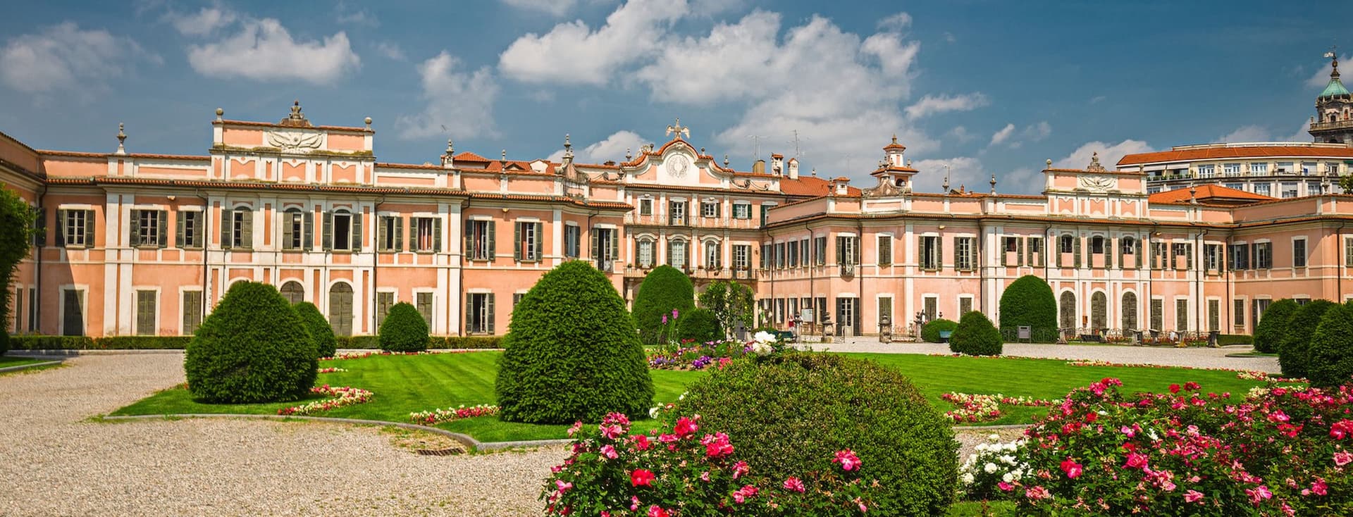 A grand peach-colored baroque palace with ornate architectural details and symmetrical wings, viewed across meticulously landscaped formal gardens featuring shaped topiary bushes, vibrant pink rose beds, green lawns, and gravel pathways, all set against a bright blue sky with scattered white clouds.