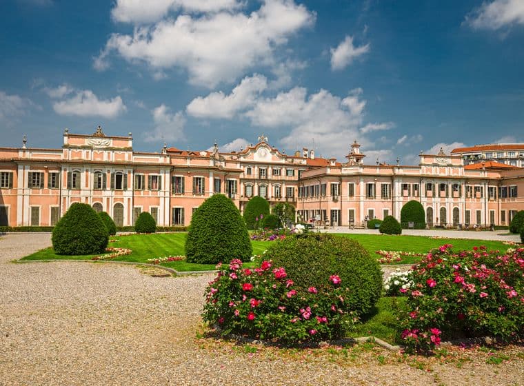 Ein großartiger pfirsichfarbener Barockpalast mit kunstvollen architektonischen Details und symmetrischen Flügeln, der über akkurat gestaltete formale Gärten mit geformten Buchsbaumhecken, leuchtenden rosa Rosenbeeten, grünen Rasenflächen und Kieswegen zu sehen ist, alles vor einem strahlend blauen Himmel mit vereinzelten weißen Wolken.