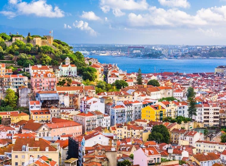 Lisbon, Portugal cityscape with colorful buildings and red tile roofs. The Tagus River and São Jorge Castle are in the background.