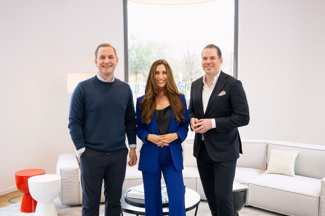 Three people smiling, standing in a modern living room with a large window, white sofa, and red and white side tables.
