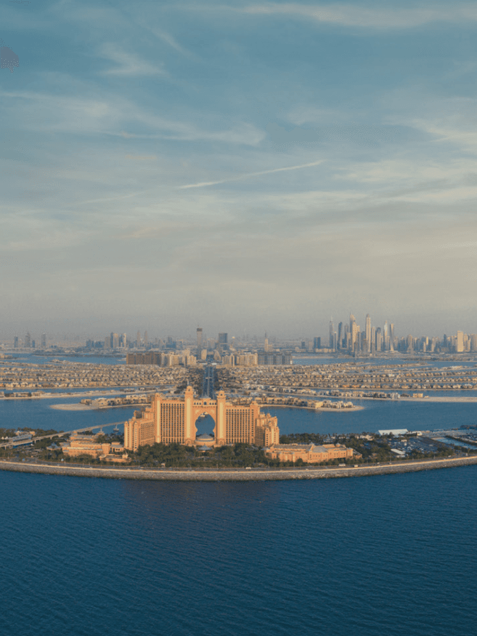 Luftaufnahme der Palm Jumeirah in Dubai, mit dem Atlantis Hotel im Vordergrund und der Skyline der Stadt im Hintergrund unter blauem Himmel.