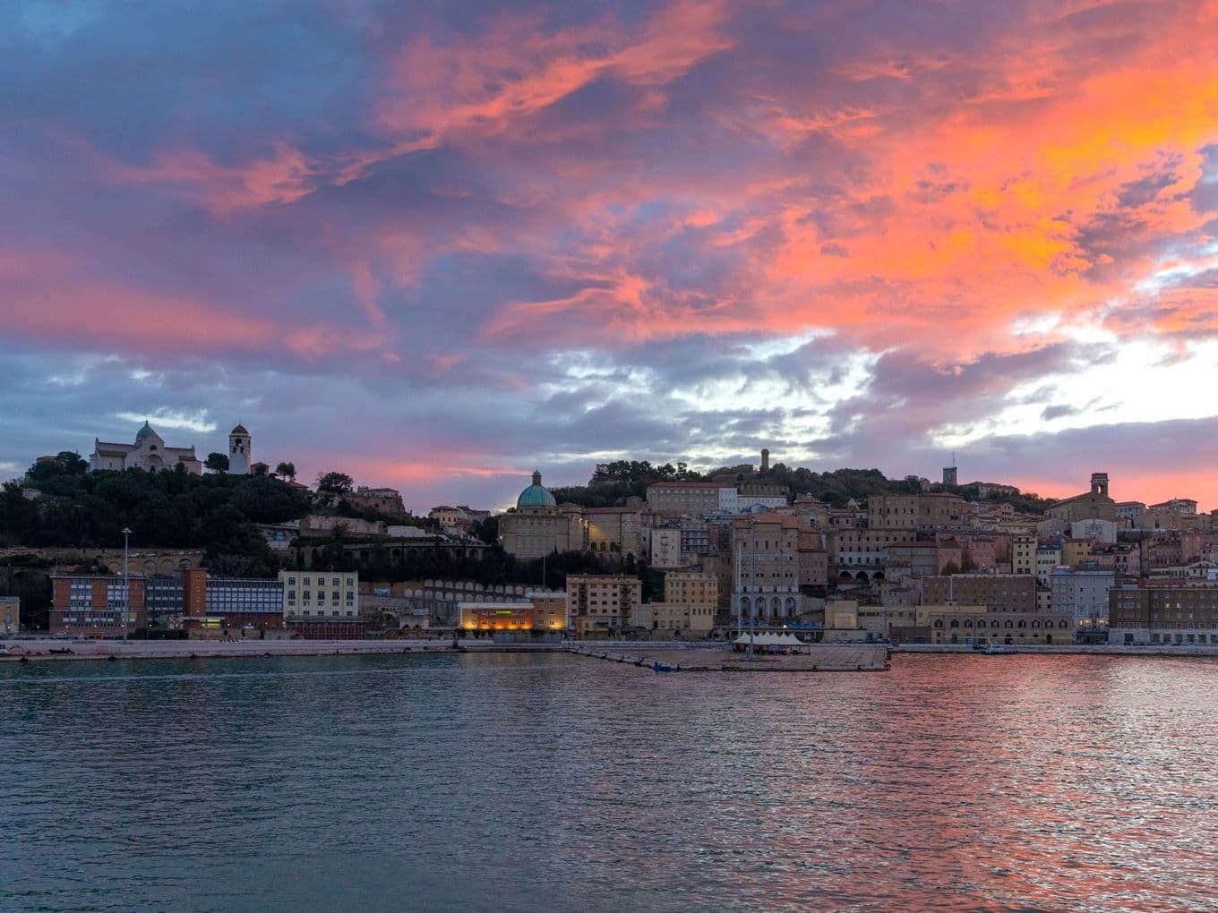 A coastal cityscape at sunset, with vibrant pink and purple clouds reflecting over calm waters, and historic buildings silhouetted against the sky.