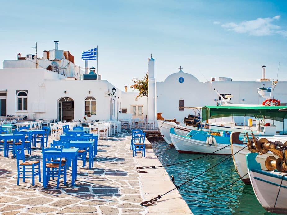Greek island scene with blue chairs and tables on a stone patio, white buildings, and fishing boats in turquoise water.