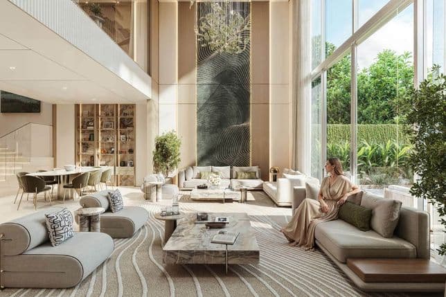 Spacious modern living room with large windows, elegant sofas, a marble table, and a woman seated near greenery, under a high ceiling chandelier.