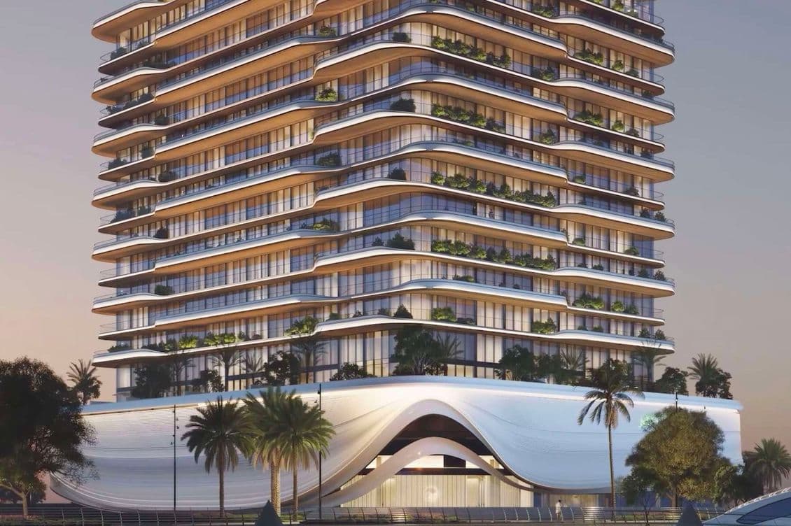 Modern waterfront building with curved balconies, illuminated at dusk. Palm trees line the entrance, and a yacht is docked nearby.