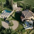Aerial view of a Tuscan villa with a terracotta roof, pool, circular driveway, and lush gardens, surrounded by vineyards and trees.