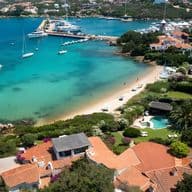 Aerial view of a scenic coastal village with turquoise waters, white sand beach, yachts, and lush greenery surrounding charming red-roofed houses.