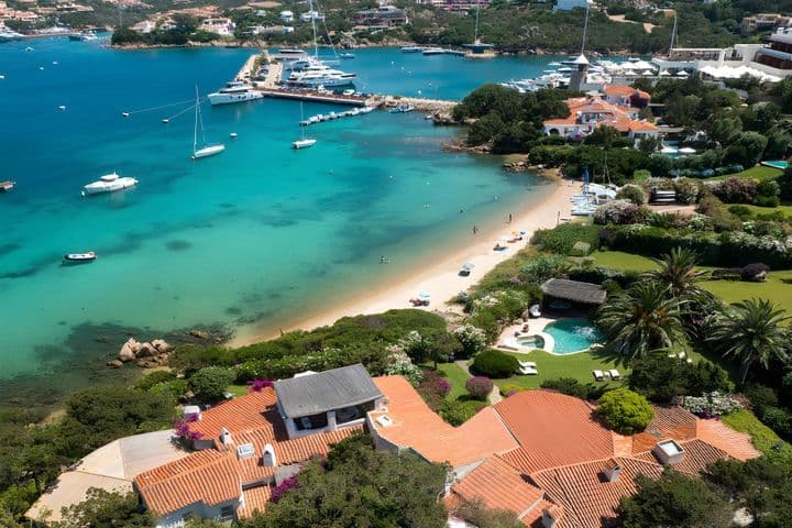 Aerial view of a scenic coastal village with turquoise waters, white sand beach, yachts, and lush greenery surrounding charming red-roofed houses.