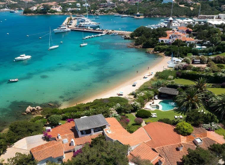 Aerial view of a scenic coastal village with turquoise waters, white sand beach, yachts, and lush greenery surrounding charming red-roofed houses.