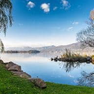 Eine friedvolle Seelandschaft Eine vollkommen ruhige Seeszene mit spiegelglatter Wasserfläche, die den tiefblauen Himmel und vereinzelte Wolken perfekt reflektiert. Im Vordergrund erstreckt sich ein grasiger Uferstreifen, gesäumt von Felsen. Bäume mit herbstlichem Laub rahmen das Bild: Eine Trauerweide auf der linken Seite und ein Baum mit goldenen Blättern auf der rechten Seite. Der spiegelglatte See erstreckt sich bis zu fernen Bergen, wobei eine kleine Insel im mittleren Hintergrund sichtbar ist. Das ruhige Wasser erzeugt perfekte Spiegelbilder der umliegenden Landschaft und schafft eine symmetrische Komposition, getaucht in weiches Morgen- oder Abendlicht.
