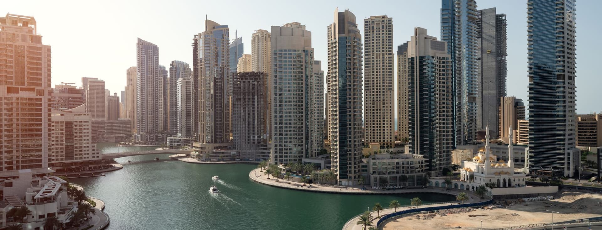 Aerial view of a modern cityscape with tall skyscrapers, a winding water canal, boats, and a road curving along the waterfront.