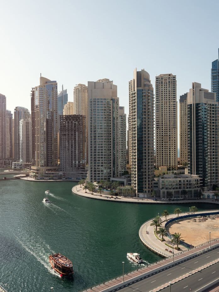 Aerial view of a modern cityscape with tall skyscrapers, a winding water canal, boats, and a road curving along the waterfront.