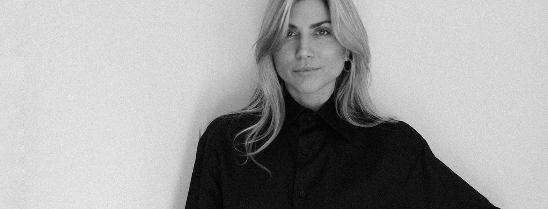 Ασπρόμαυρο πορτρέτο μιας ξανθιάς γυναίκας με μαύρο πουκάμισο με κουμπιά μπροστά σε έναν λευκό τοίχο.