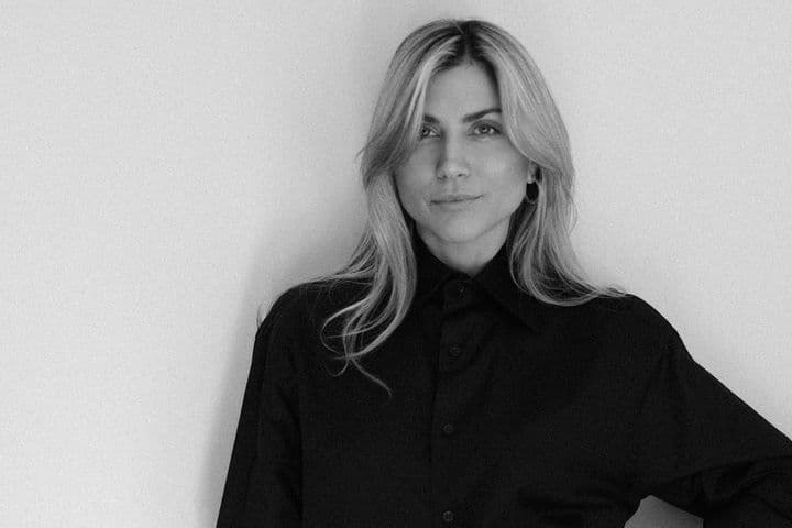 Ασπρόμαυρο πορτρέτο μιας ξανθιάς γυναίκας με μαύρο πουκάμισο με κουμπιά μπροστά σε έναν λευκό τοίχο.