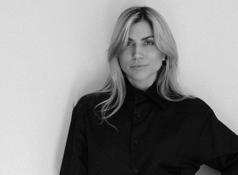 Ασπρόμαυρο πορτρέτο μιας ξανθιάς γυναίκας με μαύρο πουκάμισο με κουμπιά μπροστά σε έναν λευκό τοίχο.