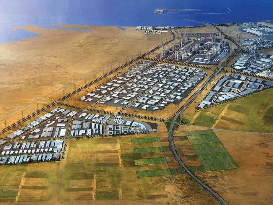 Aerial view of a structured industrial complex near a coastline, with various buildings and grid-like plots on arid land, adjacent to water.
