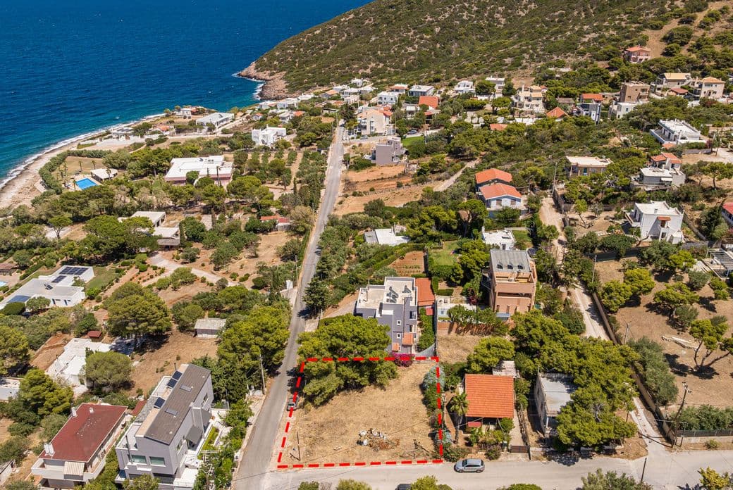 Αεροφωτογραφία μιας παραλιακής κατοικημένης περιοχής με διάσπαρτα σπίτια, καταπράσινα δέντρα και έναν δρόμο που οδηγεί στη θάλασσα. Ένα οικόπεδο σε πρώτο πλάνο περιγράφεται με κόκκινο χρώμα, τονίζοντας τα όριά του.