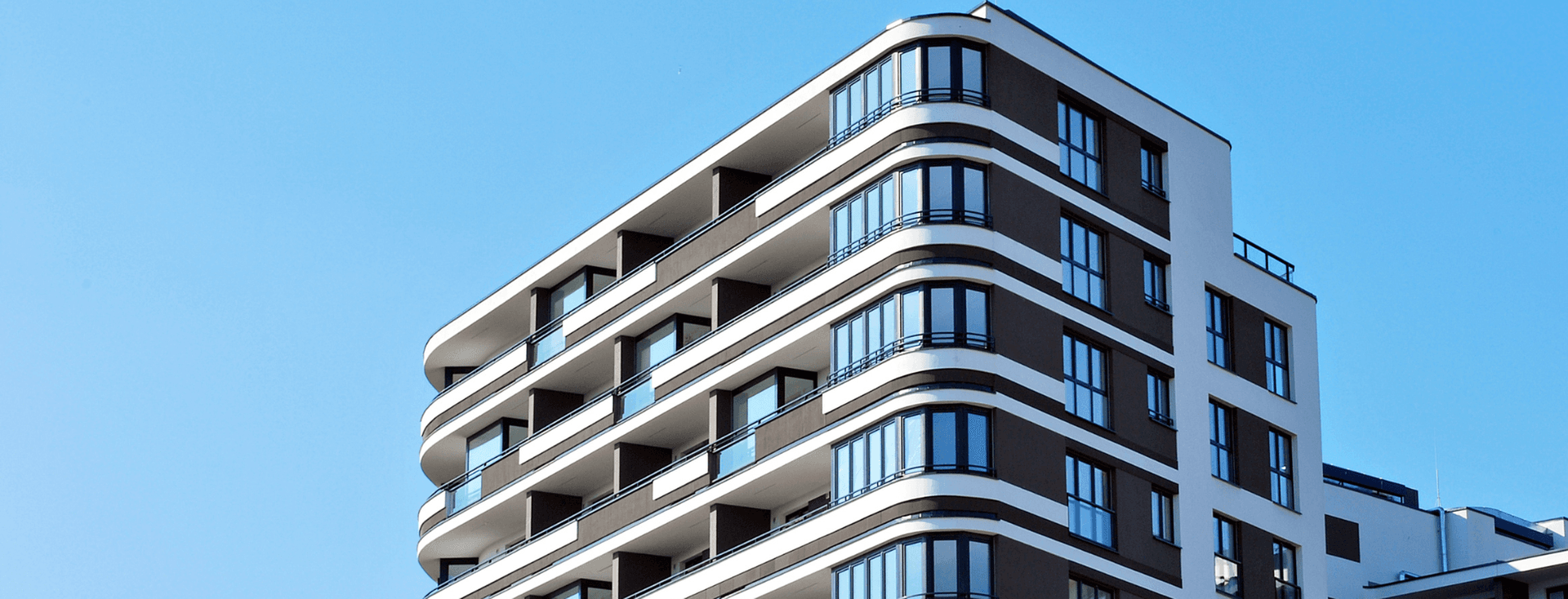 Prédio de apartamentos moderno com varandas curvas contra um céu azul claro. O prédio é marrom e branco.