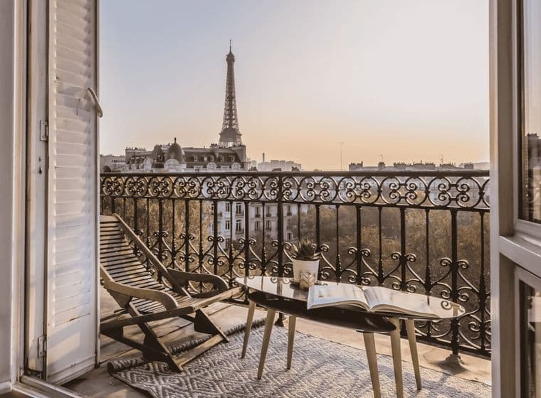Balcony view of the Eiffel Tower at sunset, with a lounge chair and table holding a book and plant, framed by open French doors.