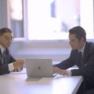 Dois homens de terno sentam-se a uma mesa branca, um usando um laptop, o outro apontando para papéis. Uma janela está no fundo.