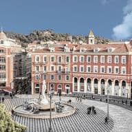 A scenic view of a European plaza with a central fountain, surrounded by red buildings with arched windows and a patterned circular pavement.