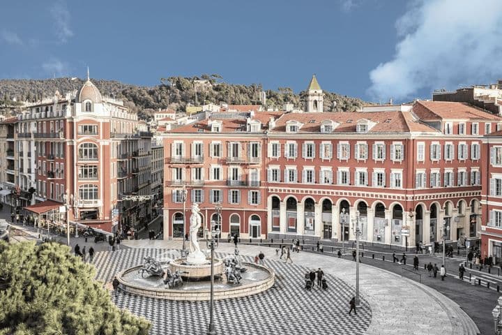 A scenic view of a European plaza with a central fountain, surrounded by red buildings with arched windows and a patterned circular pavement.