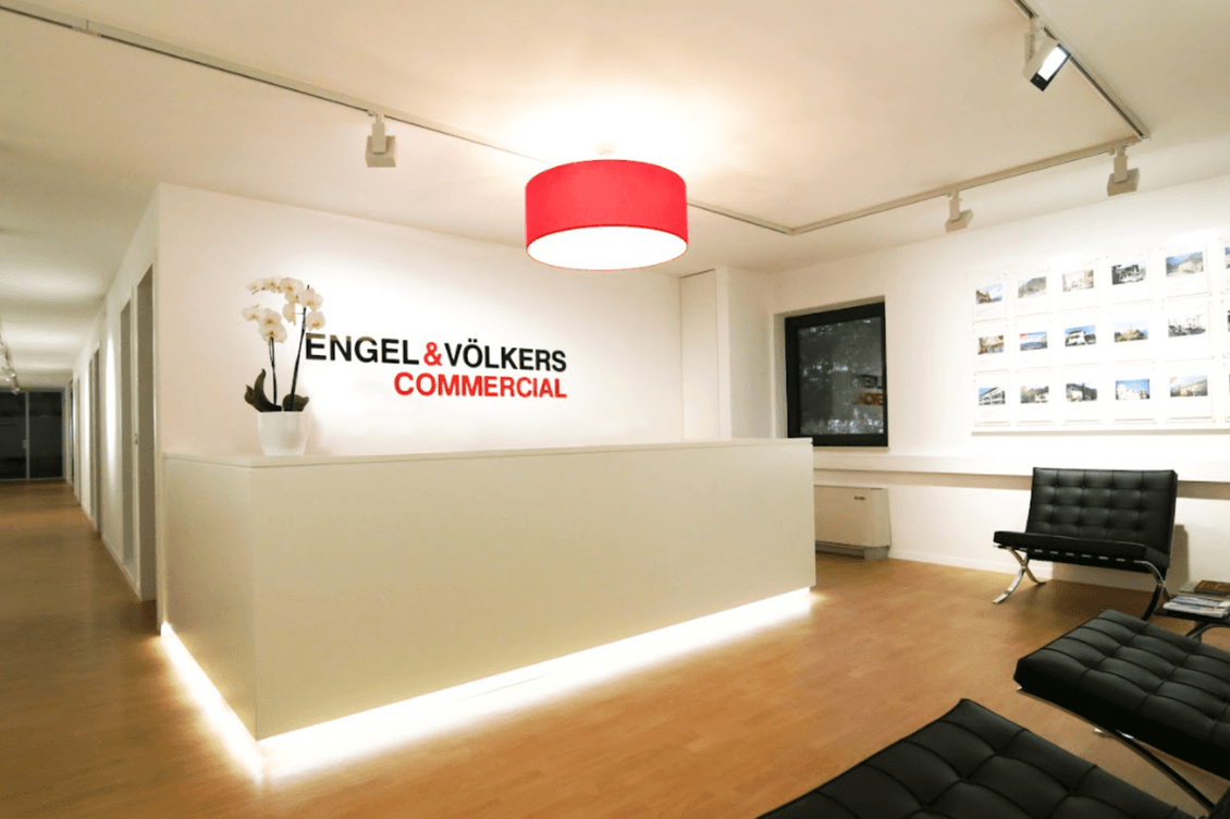 Moderner Empfangsbereich mit weißem Schreibtisch, rotem Kronleuchter und Stühlen. An den Wänden prangt das Logo von „Engel & Völkers Commercial“ sowie gerahmte Fotografien.