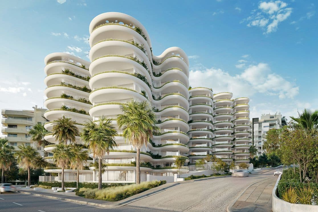Modern white apartment complex with flowing rounded balconies, planted terraces and palm trees along a sunlit urban street.
