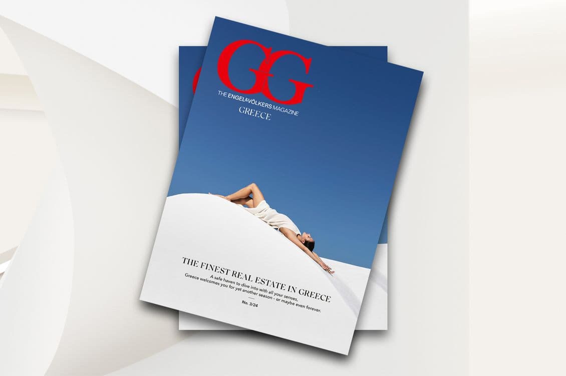 Magazine cover featuring a person lounging on a white surface against a clear blue sky. The title reads The Finest Real Estate in Greece. Bold red letters GG adorn the top. Text highlights luxury Greek properties.