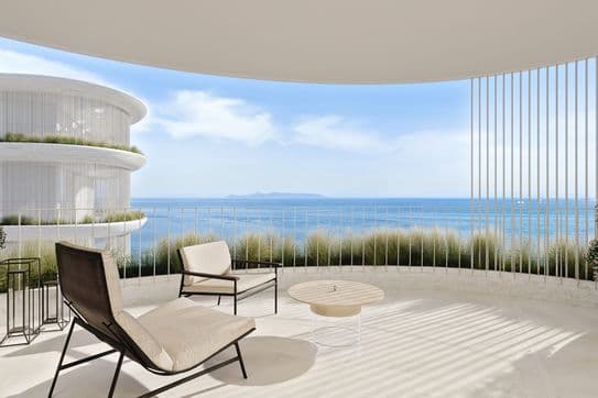 Sunlit curved balcony with lounge chairs, round table and planters overlooking blue ocean and islands through slim vertical railings.