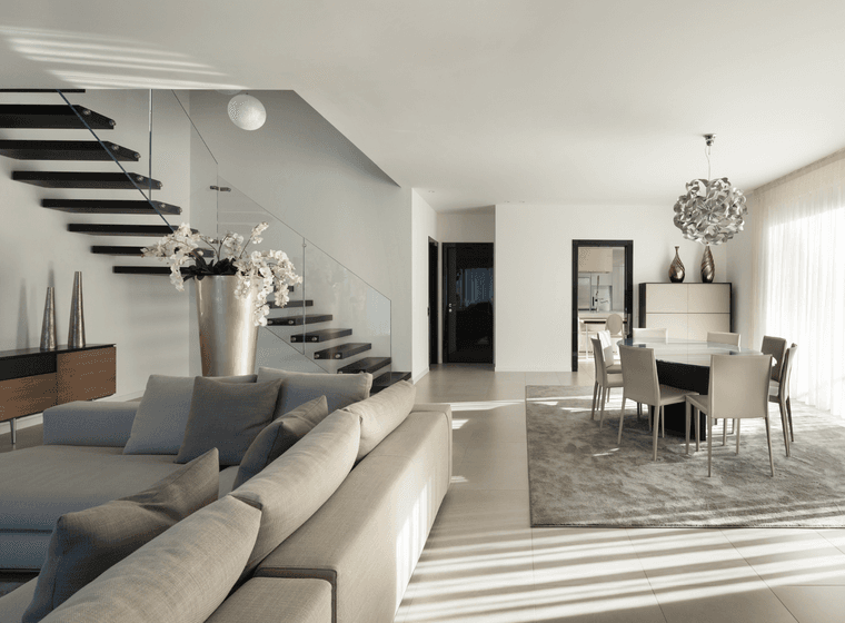 Helles, modernes Wohnzimmer mit einem grauen Sofa, einer freitragenden Treppe und einem Esstisch für sechs Personen. Sonnenlicht strömt durch zarte Vorhänge.