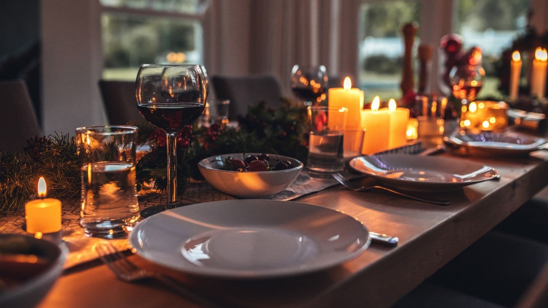 Una taula de menjador il·luminada càlidament, preparada per a un àpat festiu, amb copes de vi, plats, espelmes i decoració verda festiva.