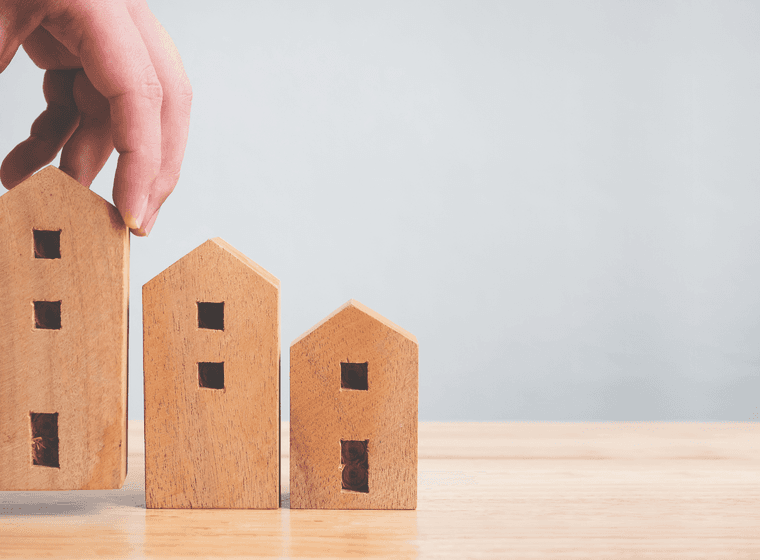 Hand placing a wooden house model next to two others on a wooden surface against a light gray background.