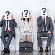 Quatre persones seuen en cadires blanques, sostenint signes d'interrogació sobre les seves cares, esperant una entrevista.