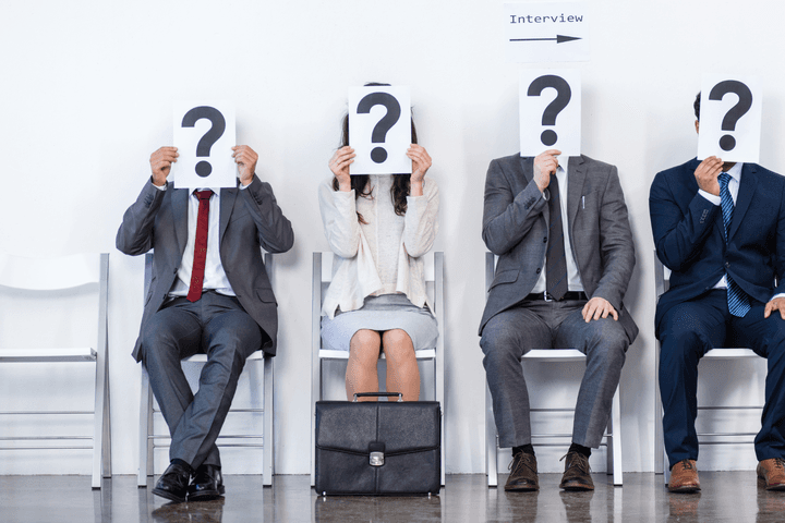 Vier Personen sitzen auf weißen Stühlen, halten Fragezeichen über ihre Gesichter und warten auf ein Interview.