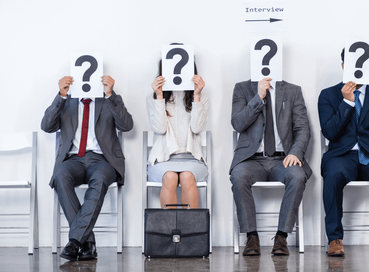 Quatre persones seuen en cadires blanques, sostenint signes d'interrogació sobre les seves cares, esperant una entrevista.