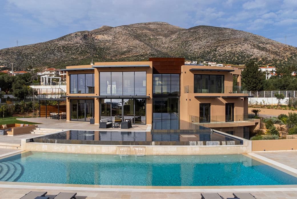 Μια μοντέρνα διώροφη βίλα με μεγάλα γυάλινα παράθυρα έχει θέα σε μια ευρύχωρη πισίνα, με φόντο καταπράσινους λόφους και διάσπαρτα σπίτια.
