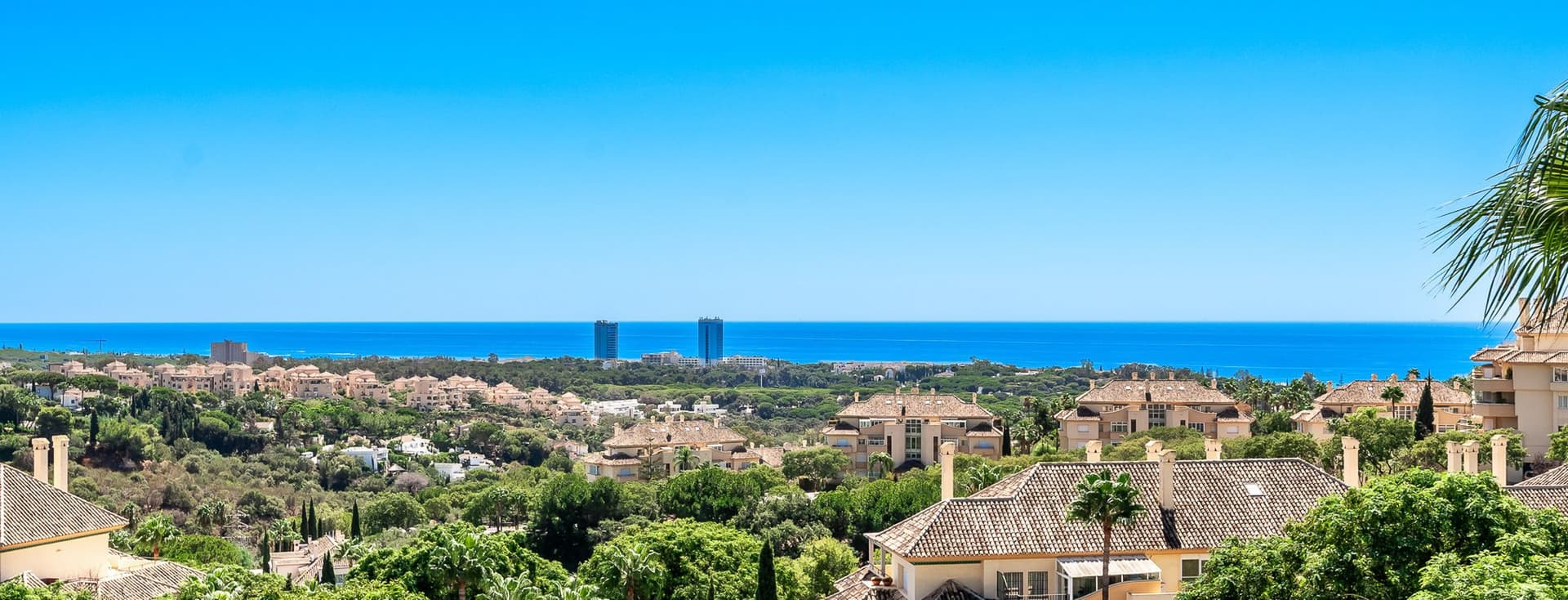 Ausblicke von Elviria aus einem Penthouse, von wo aus man viele Häuser und grüne Bäume sehen kann, im Hintergrund das Meer und den blauen Himmel.