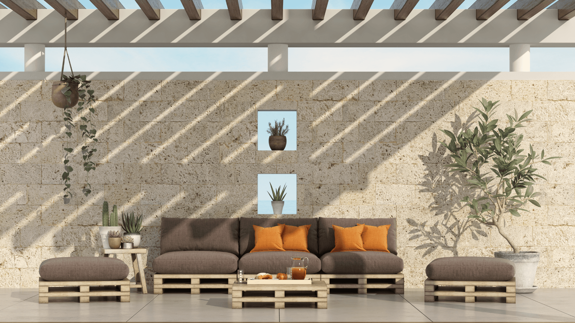 Outdoor patio with pallet furniture, brown sofa with orange pillows, and a wooden table with a pitcher of juice. Stone wall and pergola.