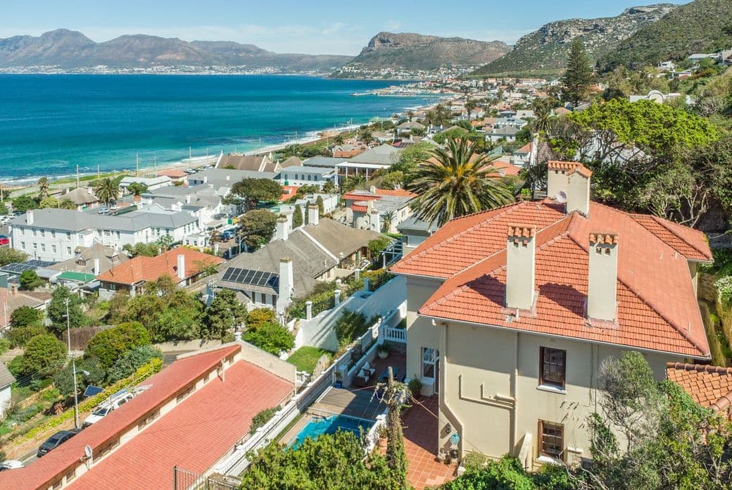 Aerial view of Kalk Bay, South Africa, showing a coastal town with terracotta-roofed houses nestled between mountains and the blue ocean. A prominent house with a swimming pool overlooks the bay and surrounding residential area.