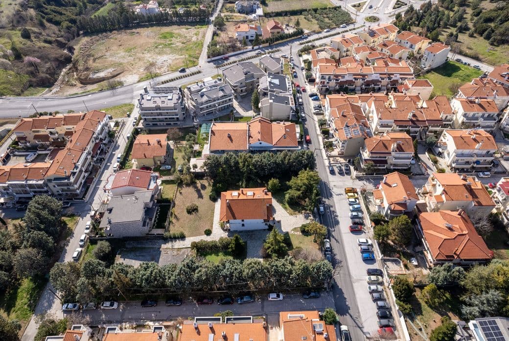 Aerial view of a residential neighborhood with red-roofed houses, tree-lined streets, and parked cars on a sunny day.