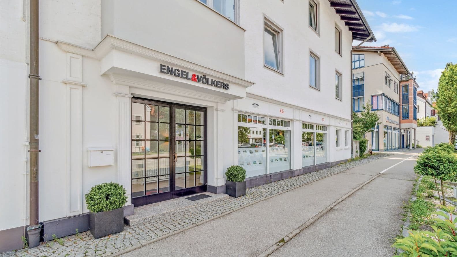 Straßenansicht eines modernen Gebäudes mit großen Fenstern und einem Schild mit der Aufschrift „Engel & Völkers“, umgeben von Grünflächen und einem gepflasterten Gehweg.