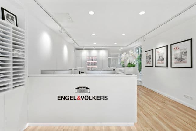 Moderner Empfangsbereich mit weißen Wänden, Holzfußboden und einer Rezeption mit dem Engel & Völkers-Logo. Gerahmte Kunst an den Wänden.