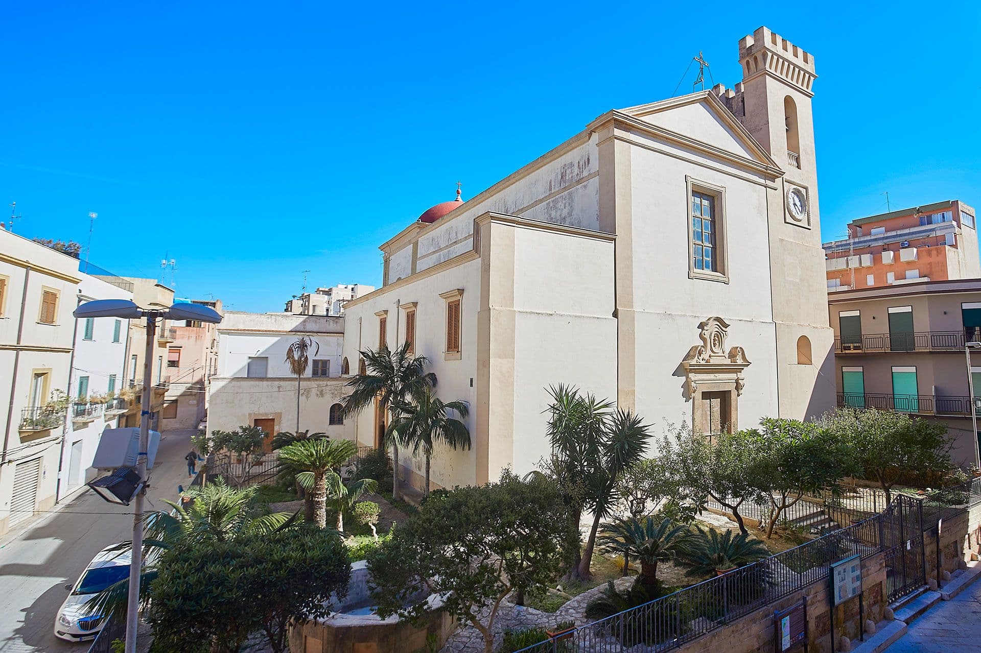 A historic church with a clock tower surrounded by palm trees and buildings under a clear blue sky.