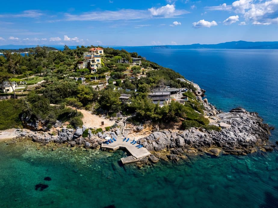 Aerial drone view of Chalkidiki’s rocky coastline with clear turquoise water, a small pier with sunbeds, lush green trees, and several houses overlooking the sea on a sunny day.