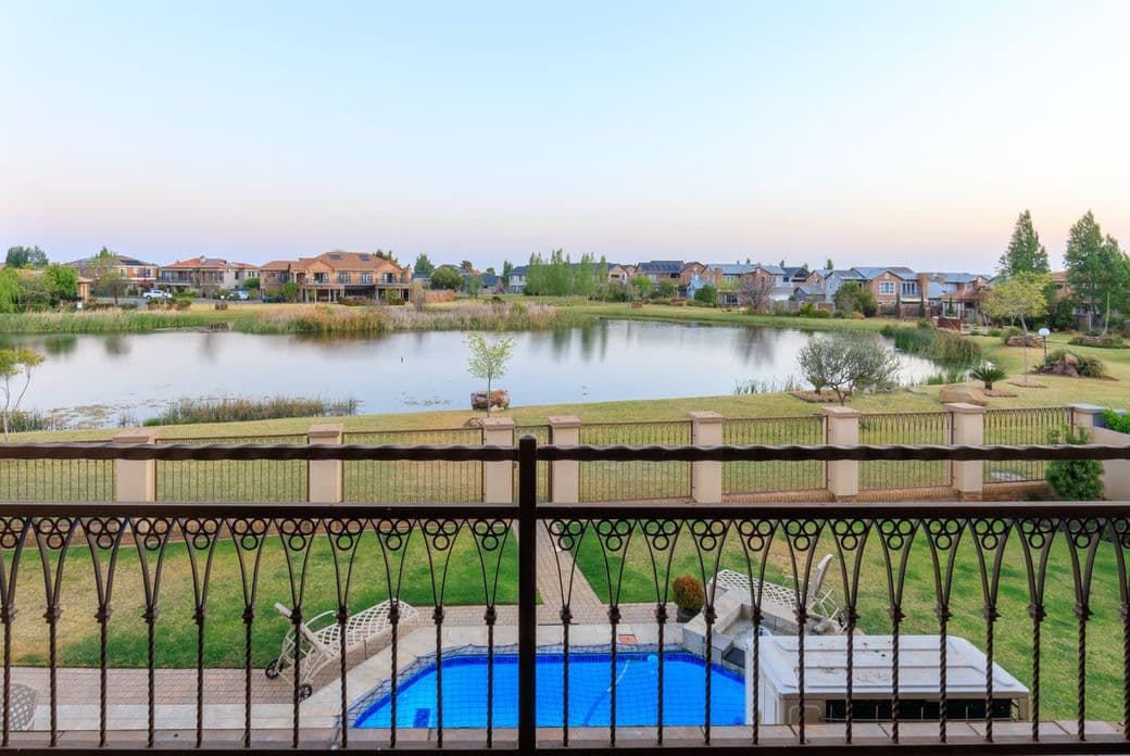 View from a balcony overlooking a residential estate in Potchefstroom, South Africa. The scene features an ornate metal railing in the foreground, a blue swimming pool below, and beyond it a small lake surrounded by green lawns and residential homes. The peaceful suburban setting is captured during early evening with a soft sky.