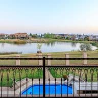 View from a balcony overlooking a residential estate in Potchefstroom, South Africa. The scene features an ornate metal railing in the foreground, a blue swimming pool below, and beyond it a small lake surrounded by green lawns and residential homes. The peaceful suburban setting is captured during early evening with a soft sky.