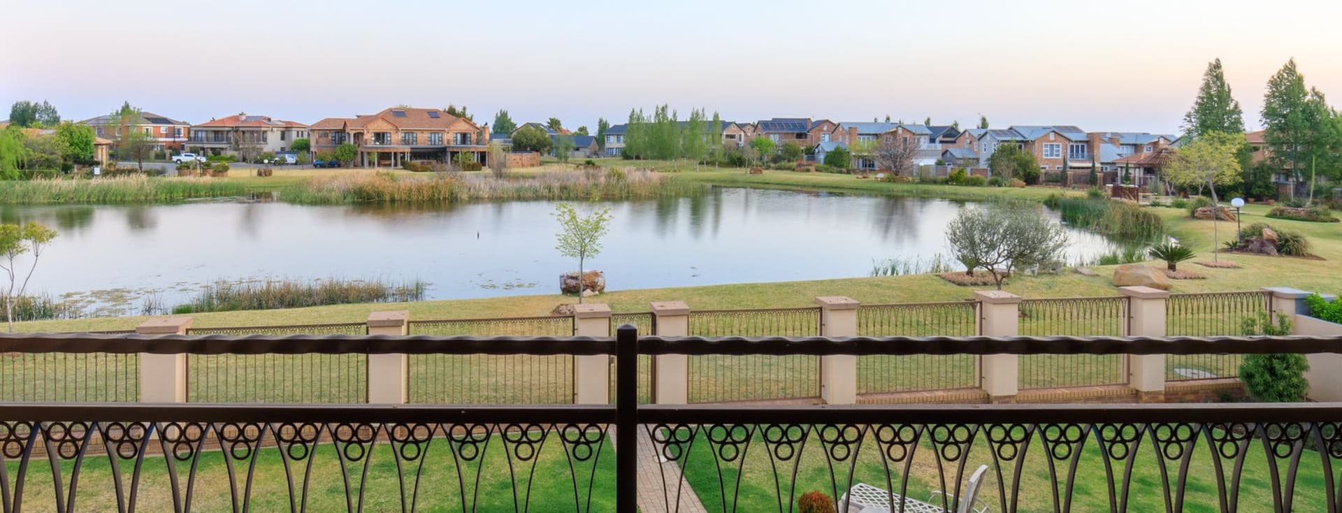 View from a balcony overlooking a residential estate in Potchefstroom, South Africa. The scene features an ornate metal railing in the foreground, a blue swimming pool below, and beyond it a small lake surrounded by green lawns and residential homes. The peaceful suburban setting is captured during early evening with a soft sky.