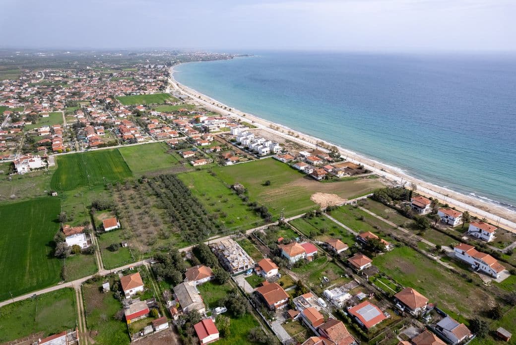 Aerial view of a coastal town with houses, green fields, and a long sandy beach next to a blue sea under a cloudy sky.